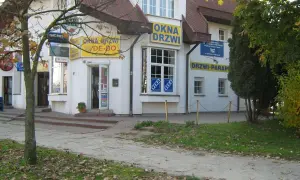 Budynek z żółtymi banerami "OKNA DRZWI" i "DRZWI PARAPETY", z drzewem i chodnikiem przed nim.