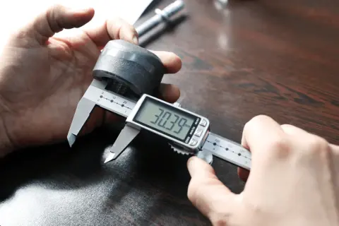 Dłoń trzymająca suwmiarkę cyfrową mierzącą metalowy element.