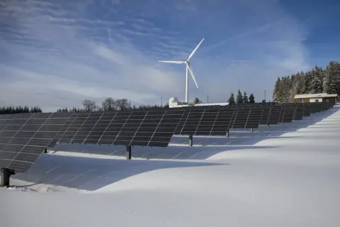 Panele słoneczne i wiatrak na ośnieżonym polu. W tle las i budynek. Taka instalacja może pomóc w stabilizacji opłata sieciowa zmienna całodobowa.