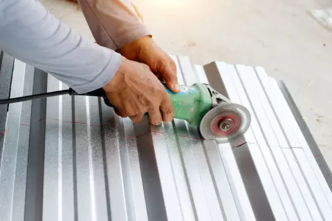 Ręce trzymają szlifierkę kątową, która przecina metalowy arkusz wzdłuż czerwonej linii.