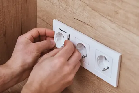 Dłonie montują białą ramkę z gniazdkami elektrycznymi na drewnianej ścianie.