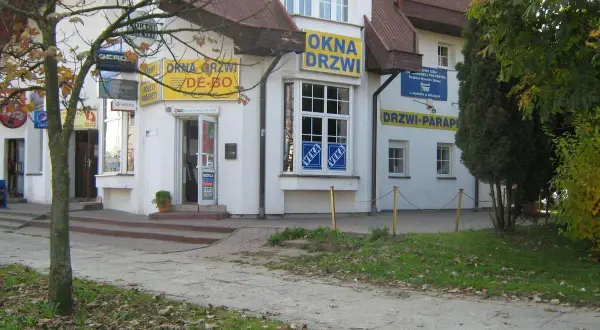 Budynek z żółtymi banerami "OKNA DRZWI" i "DRZWI PARAPETY", z drzewem i chodnikiem przed nim.