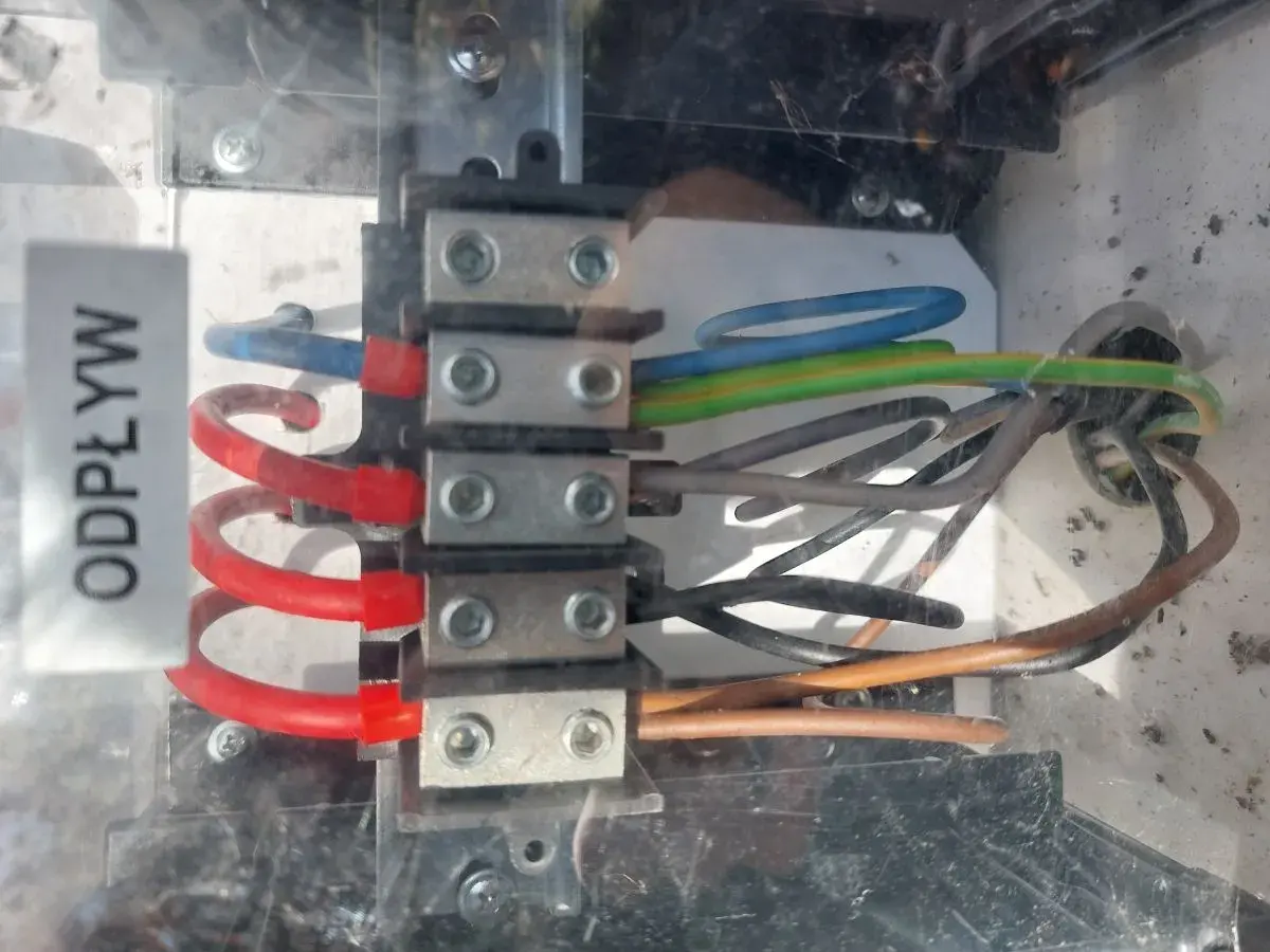 Widok z bliska na zaciski elektryczne z podłączonymi przewodami w różnych kolorach (niebieski, zielono-żółty, brązowy, czarny).