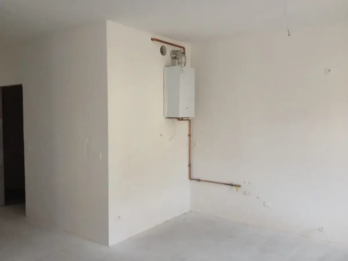 Widok na pomieszczenie, gdzie widać jak zabudować piec gazowy. Biały piec wiszący na ścianie, połączony miedzianymi rurkami z instalacją.